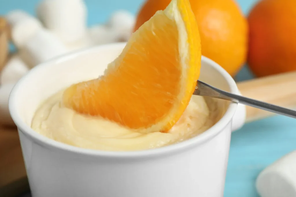 Crema all'arancia senza uova e latte in una coppetta bianca, con uno spicchio d'arancia intinto nella crema vellutata e arance intere sullo sfondo