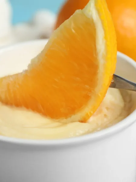 Crema all'arancia senza uova e latte in una coppetta bianca, con uno spicchio d'arancia intinto nella crema vellutata e arance intere sullo sfondo