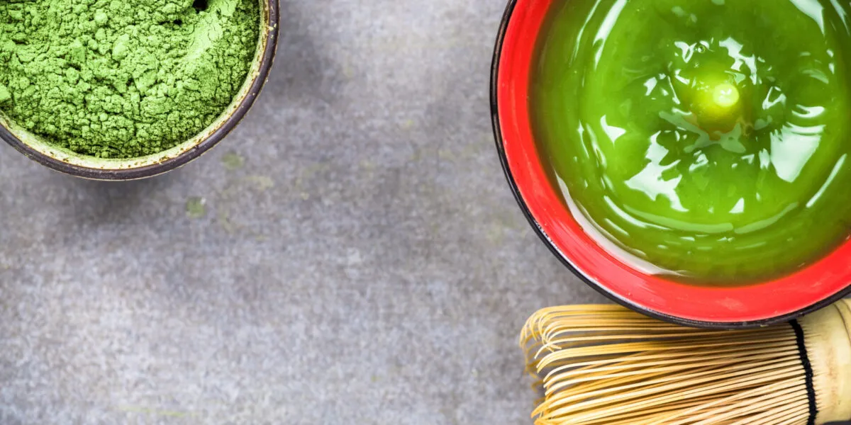 Tazza di tè Matcha verde con polvere di matcha e frustino tradizionale giapponese su sfondo grigio.