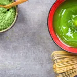 Tazza di tè Matcha verde con polvere di matcha e frustino tradizionale giapponese su sfondo grigio.