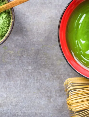 Tazza di tè Matcha verde con polvere di matcha e frustino tradizionale giapponese su sfondo grigio.