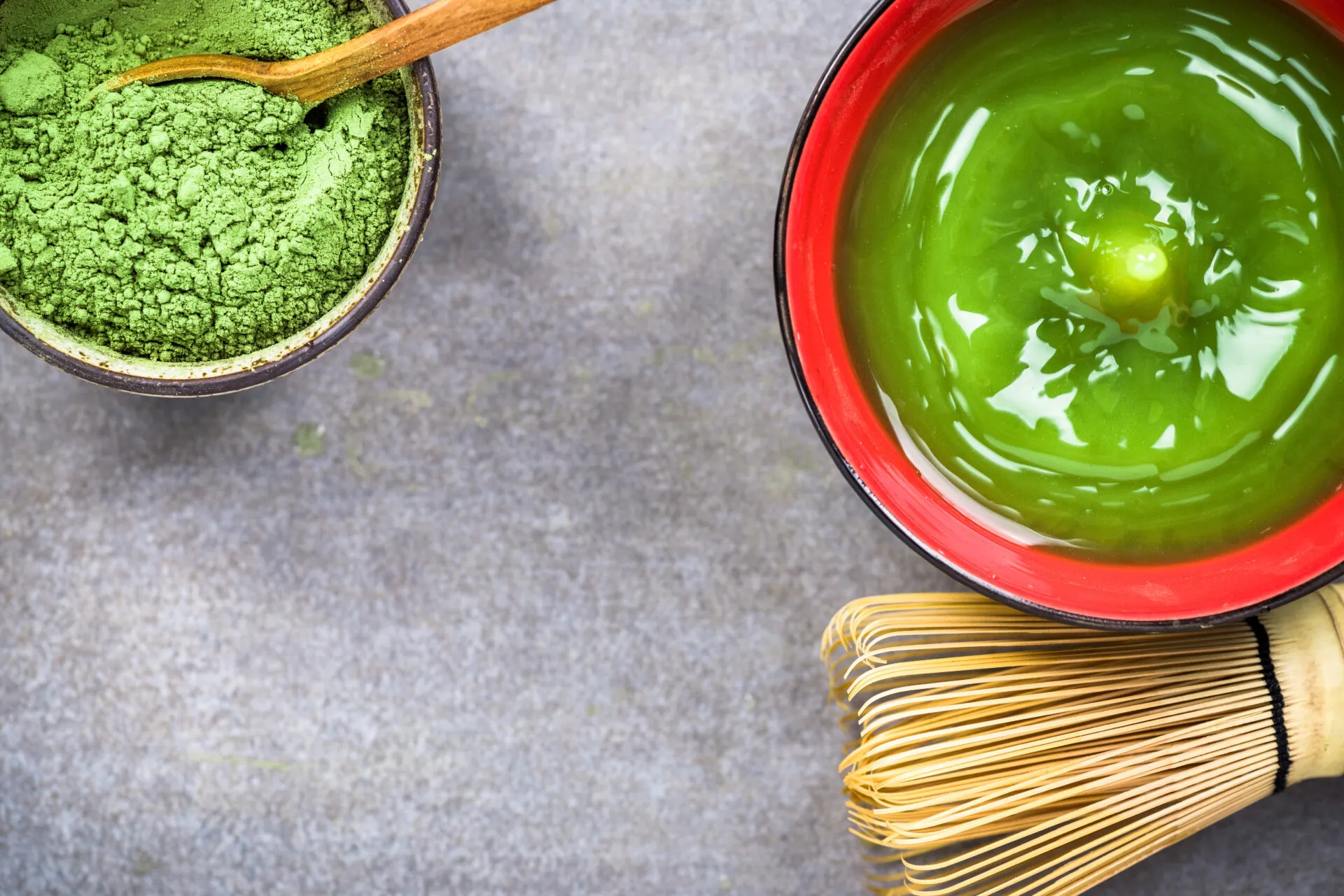 Tazza di tè Matcha verde con polvere di matcha e frustino tradizionale giapponese su sfondo grigio.