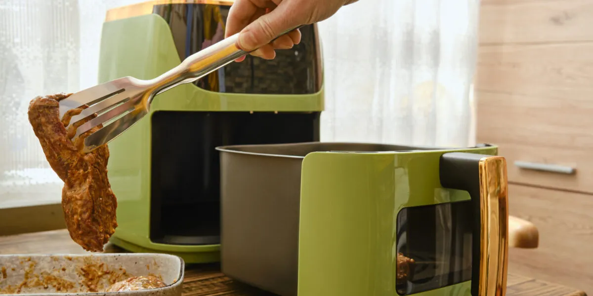 Friggitrice ad aria moderna con cestello estraibile mentre viene inserita una bistecca con una pinza da cucina.