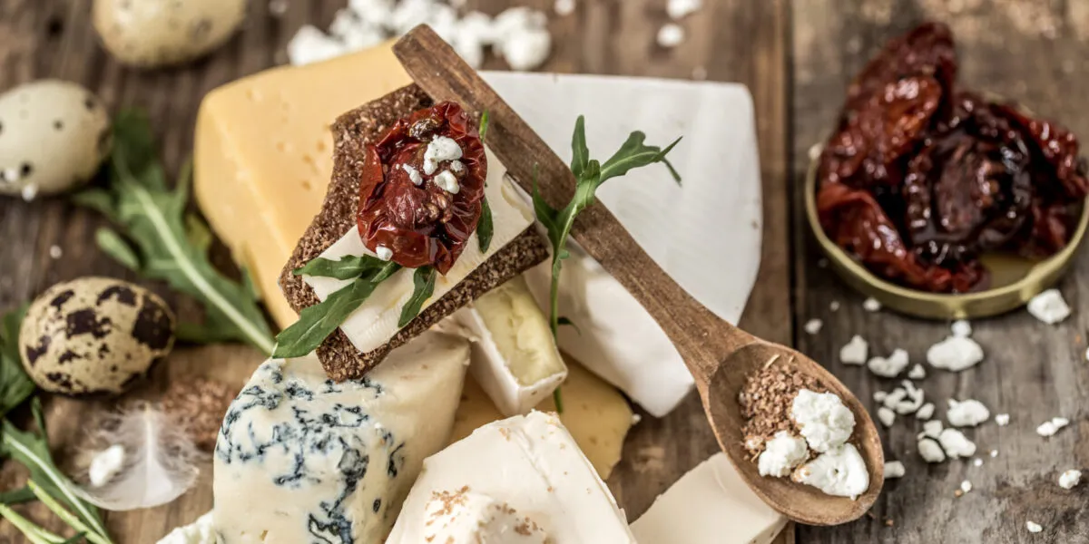Tagliere con assortimento di formaggi per antipasti freschi e stagionati, cracker croccanti, pomodori secchi e rucola su tavolo rustico.