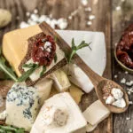 Tagliere con assortimento di formaggi per antipasti freschi e stagionati, cracker croccanti, pomodori secchi e rucola su tavolo rustico.