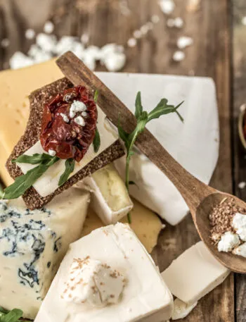 Tagliere con assortimento di formaggi per antipasti freschi e stagionati, cracker croccanti, pomodori secchi e rucola su tavolo rustico.