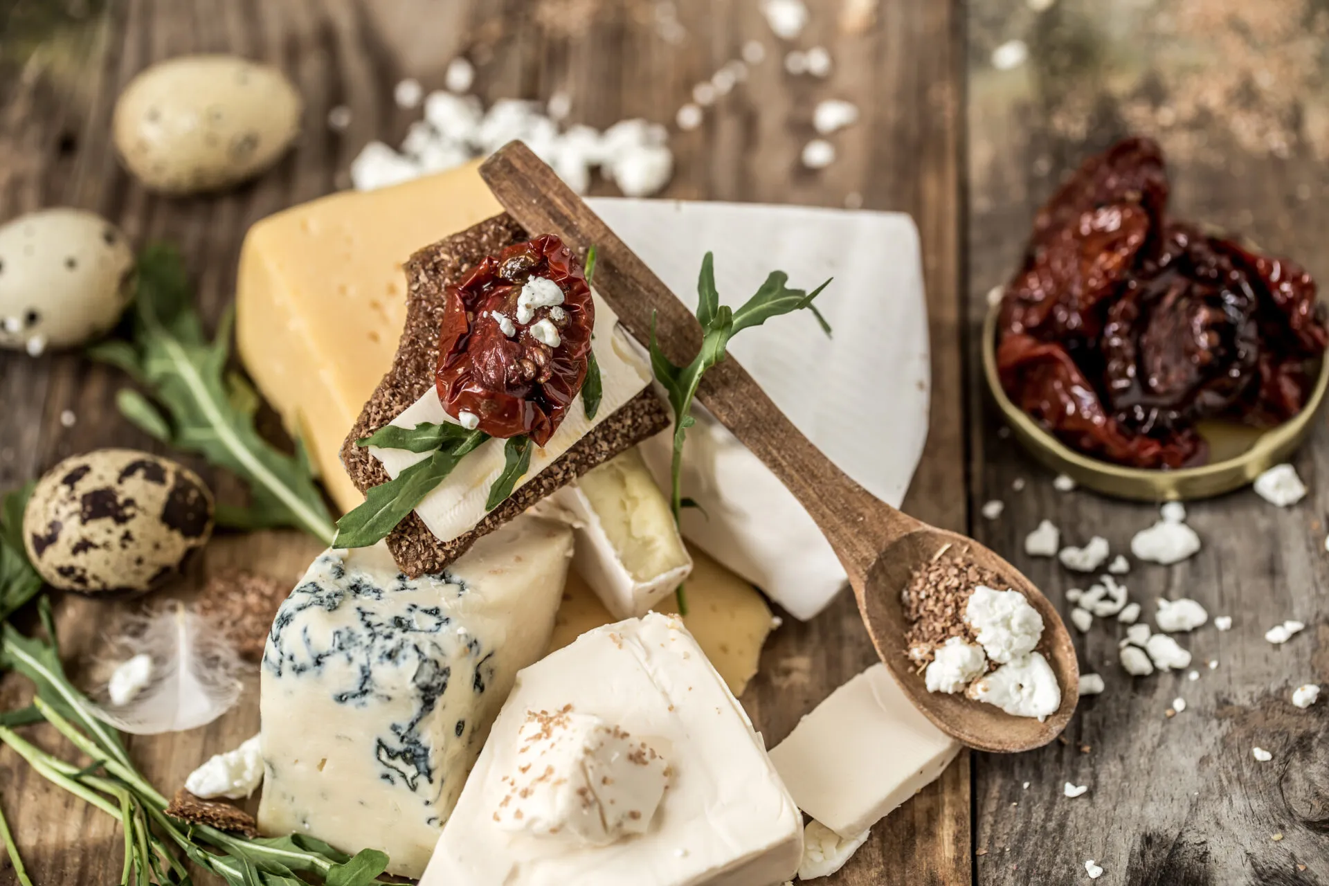 Tagliere con assortimento di formaggi per antipasti freschi e stagionati, cracker croccanti, pomodori secchi e rucola su tavolo rustico.