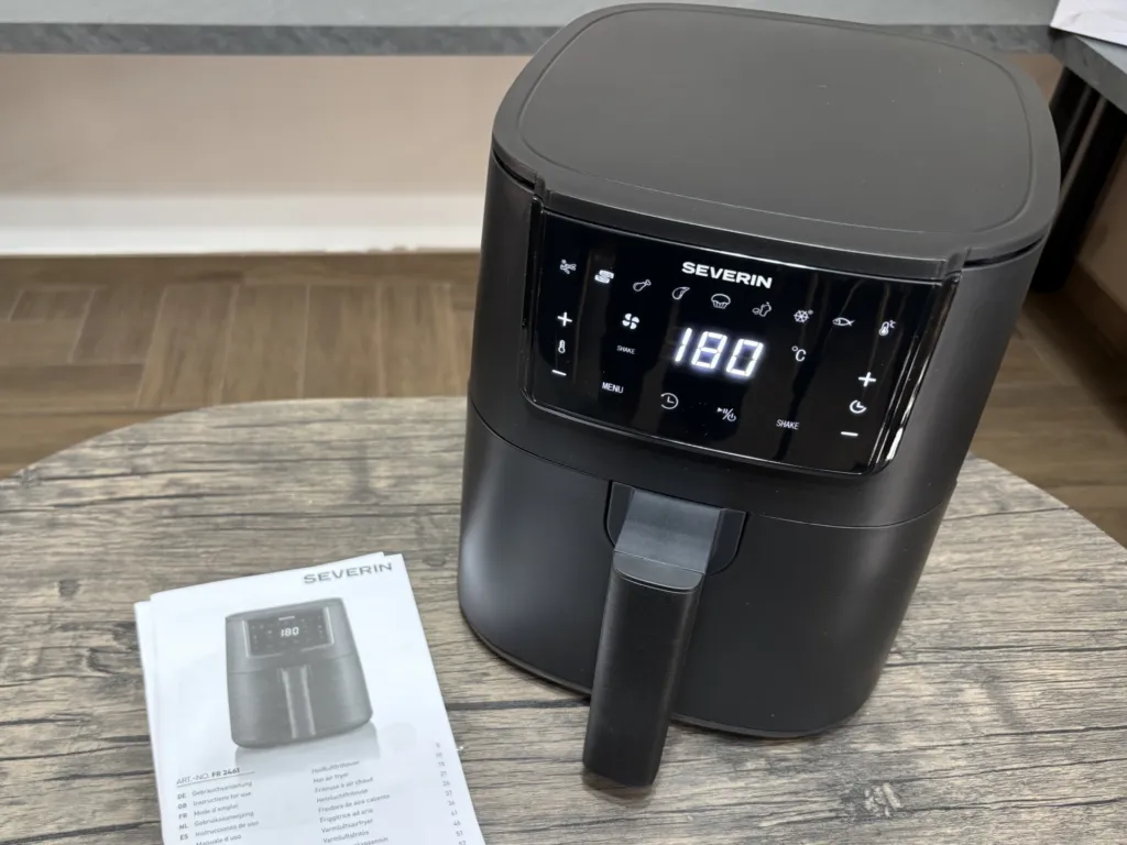 Friggitrice ad aria Severin FR 2461 Black Line posizionata su un tavolo in legno con manuale di istruzioni accanto, dotata di display digitale touch acceso a 180 °C e design compatto nero opaco.
