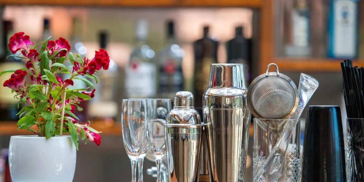 Accessori da bar per preparare cocktail analcolici estivi su un bancone, con shaker in acciaio, bicchieri e fiori decorativi sullo sfondo di bottiglie sfocate.