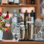 Accessori da bar per preparare cocktail analcolici estivi su un bancone, con shaker in acciaio, bicchieri e fiori decorativi sullo sfondo di bottiglie sfocate.