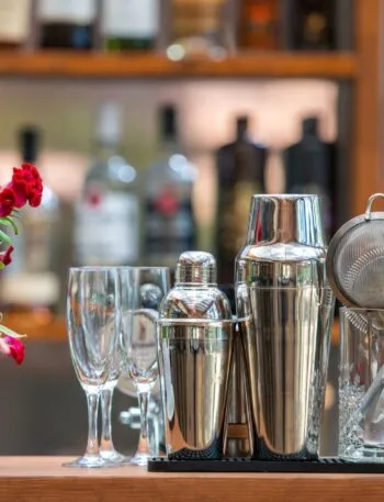 Accessori da bar per preparare cocktail analcolici estivi su un bancone, con shaker in acciaio, bicchieri e fiori decorativi sullo sfondo di bottiglie sfocate.