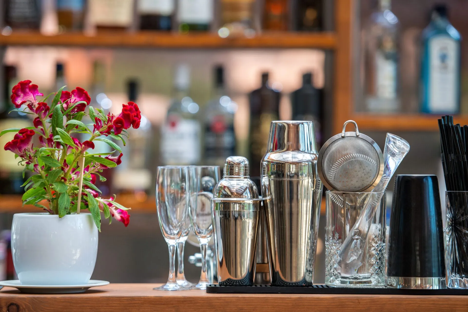 Accessori da bar per preparare cocktail analcolici estivi su un bancone, con shaker in acciaio, bicchieri e fiori decorativi sullo sfondo di bottiglie sfocate.