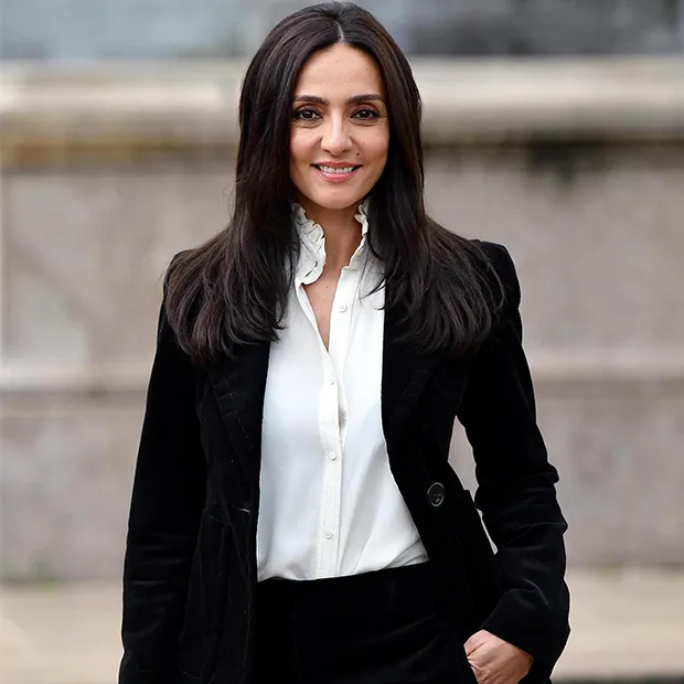 Ambra Angiolini posa all'aperto con blazer nero e camicia bianca, sorridendo verso l'obiettivo.