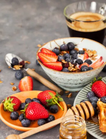 Colazione fit con yogurt, granola, fragole, mirtilli, waffle integrali e caffè per iniziare la giornata con energia e leggerezza.