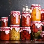 Barattoli di conserve fatte in casa con pomodori, peperoni, cetrioli e verdure sott'aceto disposti su un tavolo, esempio di preparazione sicura delle conserve domestiche.
