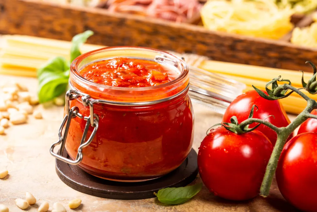 Barattolo di conserva di pomodoro fatta in casa con salsa rossa, pomodori freschi e basilico su un piano da cucina, simbolo della tradizione delle conserve domestiche.