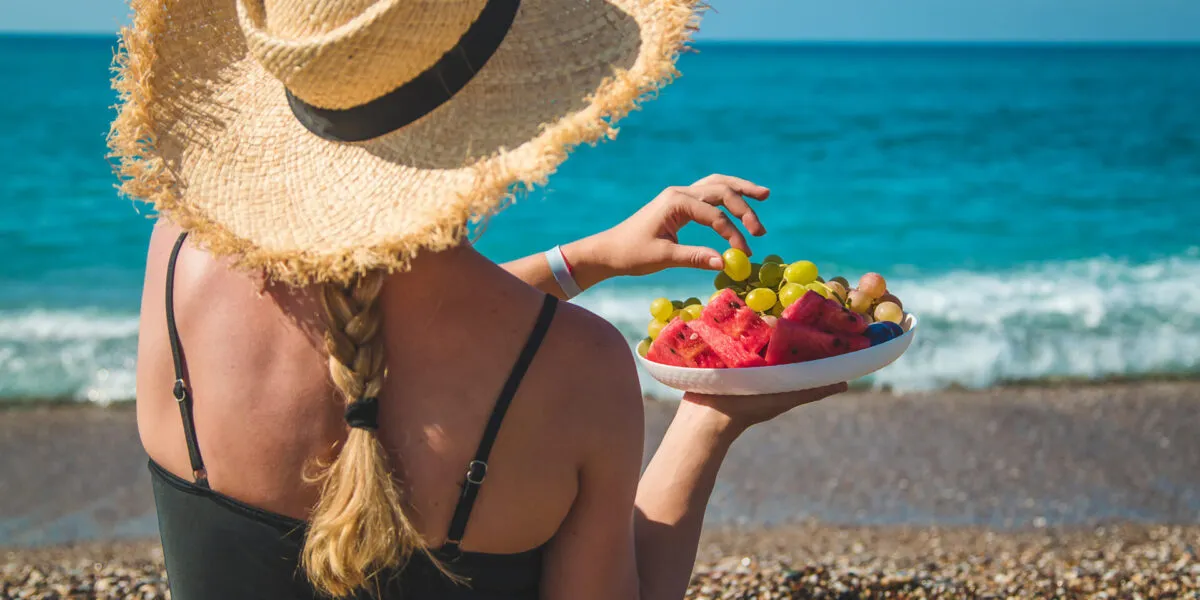 Donna in spiaggia con cappello di paglia che mangia frutta fresca davanti al mare, simbolo di un pranzo leggero e salutare da portare in spiaggia.