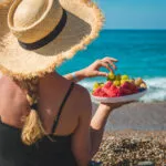 Donna in spiaggia con cappello di paglia che mangia frutta fresca davanti al mare, simbolo di un pranzo leggero e salutare da portare in spiaggia.