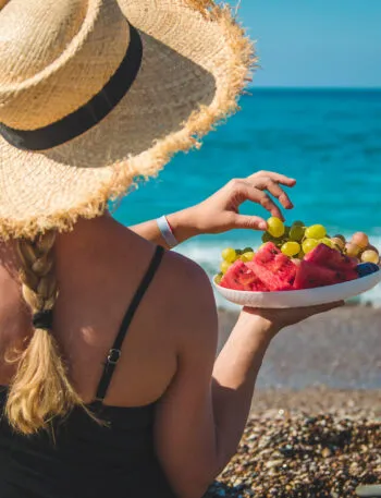 Donna in spiaggia con cappello di paglia che mangia frutta fresca davanti al mare, simbolo di un pranzo leggero e salutare da portare in spiaggia.