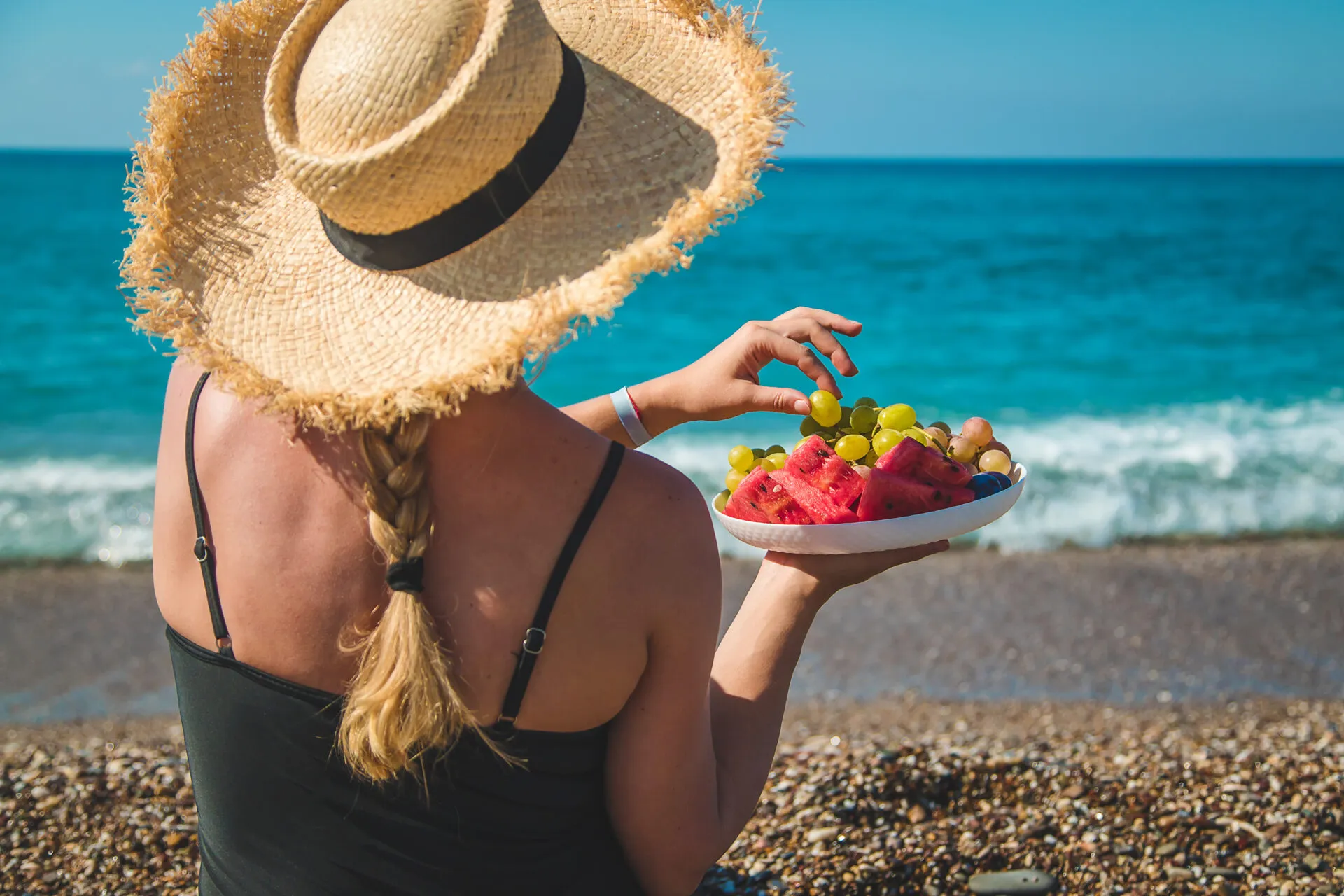 Donna in spiaggia con cappello di paglia che mangia frutta fresca davanti al mare, simbolo di un pranzo leggero e salutare da portare in spiaggia.