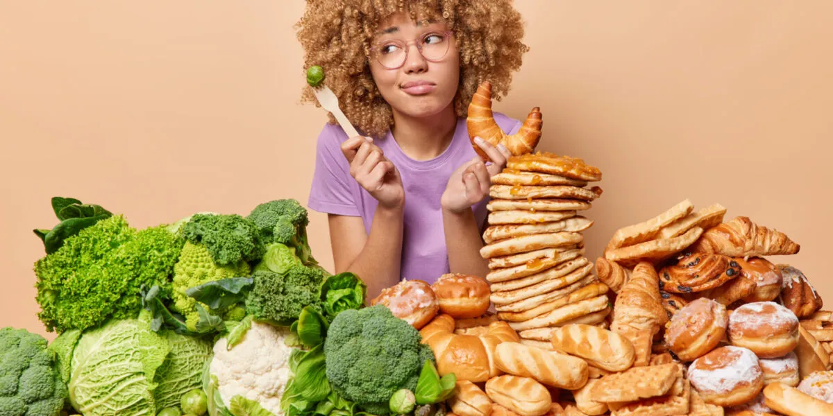 Donna indecisa tra verdure e alimenti ricchi di carboidrati come pane, croissant e pancake, simbolo della scelta di eliminare i carboidrati dalla dieta.