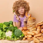 Donna indecisa tra verdure e alimenti ricchi di carboidrati come pane, croissant e pancake, simbolo della scelta di eliminare i carboidrati dalla dieta.