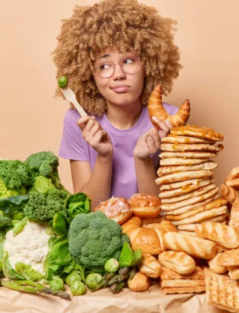 Donna indecisa tra verdure e alimenti ricchi di carboidrati come pane, croissant e pancake, simbolo della scelta di eliminare i carboidrati dalla dieta.