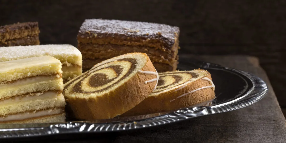 Assortimento di dolci da credenza su vassoio argentato: rotolo bicolore, torta a strati e quadrotti soffici, esempi di dolci che si conservano per giorni senza frigorifero.