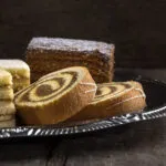 Assortimento di dolci da credenza su vassoio argentato: rotolo bicolore, torta a strati e quadrotti soffici, esempi di dolci che si conservano per giorni senza frigorifero.
