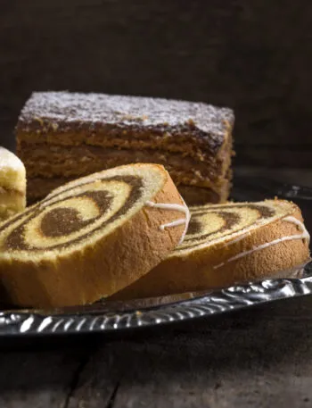 Assortimento di dolci da credenza su vassoio argentato: rotolo bicolore, torta a strati e quadrotti soffici, esempi di dolci che si conservano per giorni senza frigorifero.
