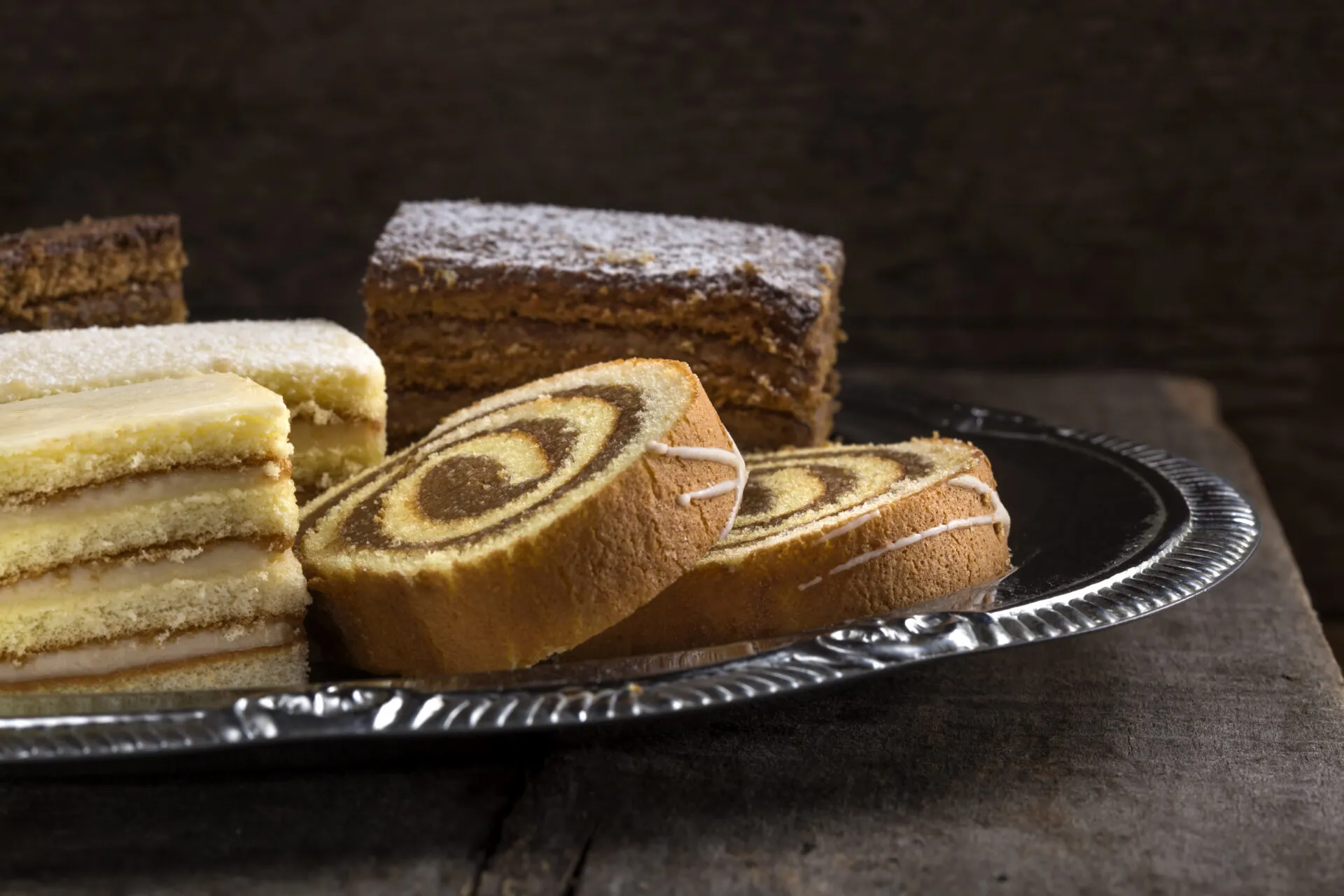 Assortimento di dolci da credenza su vassoio argentato: rotolo bicolore, torta a strati e quadrotti soffici, esempi di dolci che si conservano per giorni senza frigorifero.