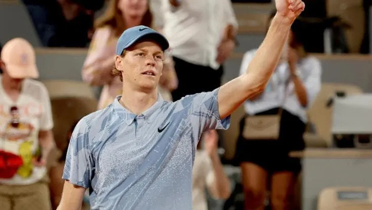 Jannik Sinner saluta il pubblico sul campo da tennis al termine di un incontro, con il braccio alzato e abbigliamento sportivo.