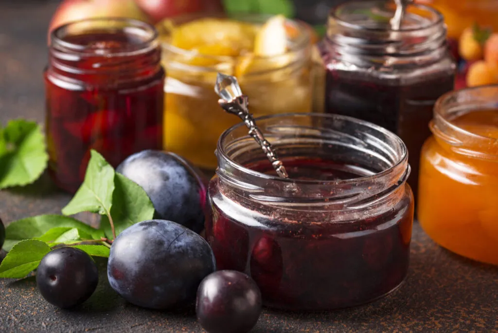 Vasetti di marmellata fatta in casa ai frutti estivi, con confettura di prugne, albicocche e frutta fresca disposti su un tavolo rustico.