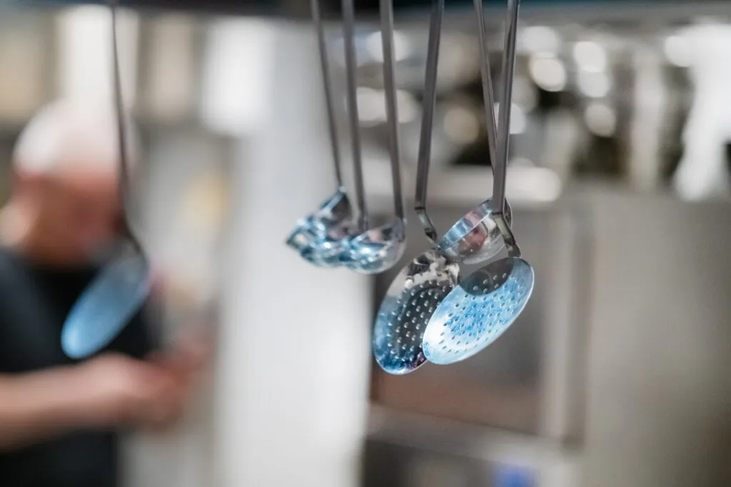 Mestoli e schiumarole in acciaio inox appesi in cucina professionale, utensili resistenti e igienici per la preparazione dei cibi.
