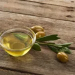 Ciotola di olio extravergine d'oliva con olive e foglie di ulivo su un tavolo in legno, simbolo di un olio EVO di qualità.