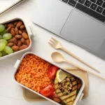 Lunch box per il pranzo in ufficio con lenticchie rosse, melanzana grigliata, pomodorini, mandorle, mela e uva, ideale per un pasto fit e sano.