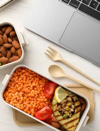 Lunch box per il pranzo in ufficio con lenticchie rosse, melanzana grigliata, pomodorini, mandorle, mela e uva, ideale per un pasto fit e sano.