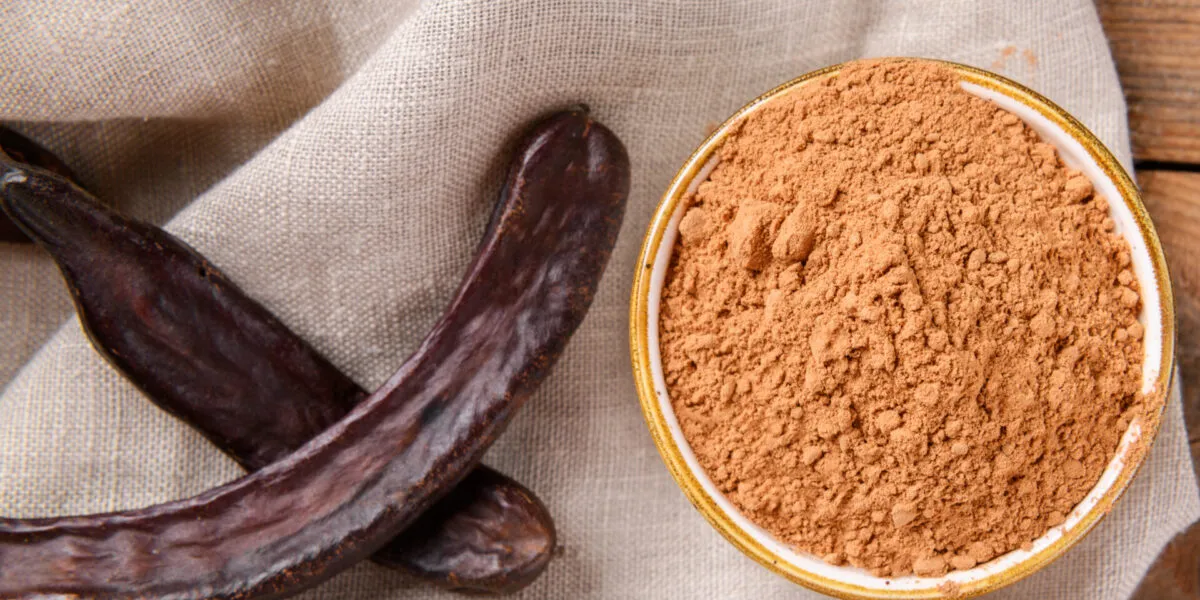 Farina di carruba in polvere all'interno di una ciotola con baccelli di carruba essiccati su tessuto naturale, ingrediente utilizzato in numerose ricette dolci e vegan.