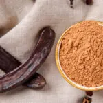 Farina di carruba in polvere all'interno di una ciotola con baccelli di carruba essiccati su tessuto naturale, ingrediente utilizzato in numerose ricette dolci e vegan.
