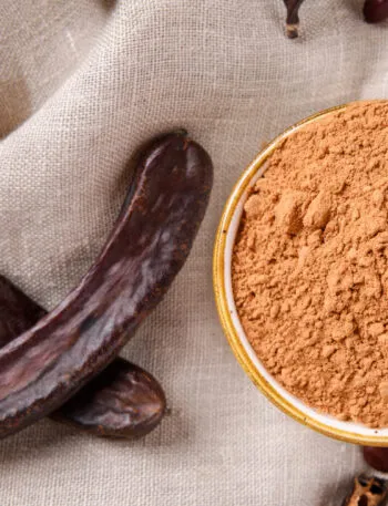 Farina di carruba in polvere all'interno di una ciotola con baccelli di carruba essiccati su tessuto naturale, ingrediente utilizzato in numerose ricette dolci e vegan.
