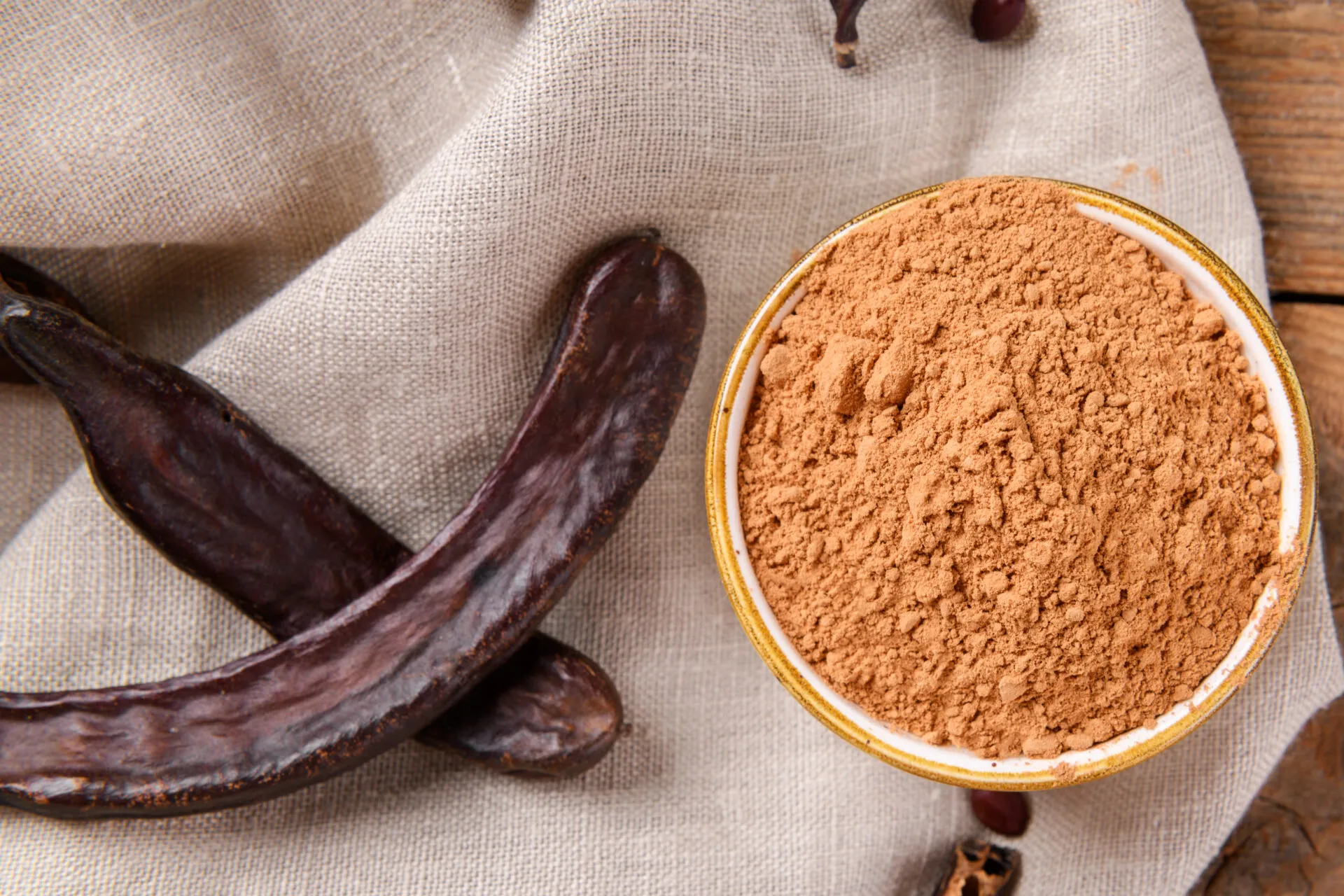 Farina di carruba in polvere all'interno di una ciotola con baccelli di carruba essiccati su tessuto naturale, ingrediente utilizzato in numerose ricette dolci e vegan.