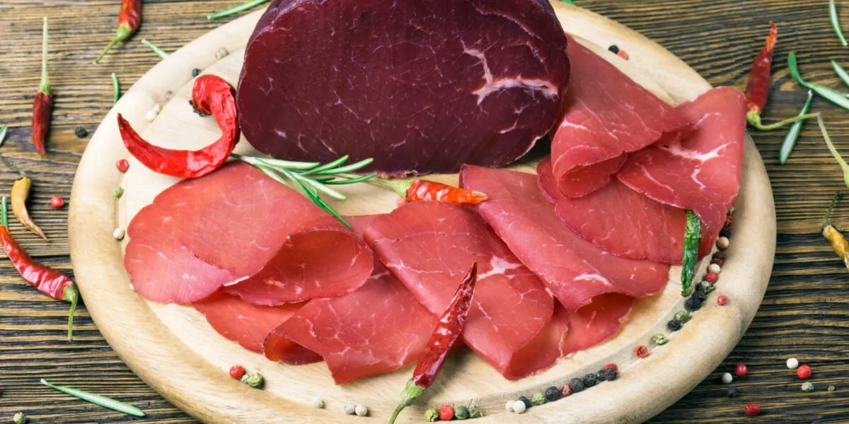 Bresaola affettata e trancio intero su tagliere in legno con rosmarino, pepe e peperoncini, ingrediente leggero e proteico ideale per ricette estive veloci e fit.