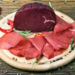 Bresaola affettata e trancio intero su tagliere in legno con rosmarino, pepe e peperoncini, ingrediente leggero e proteico ideale per ricette estive veloci e fit.
