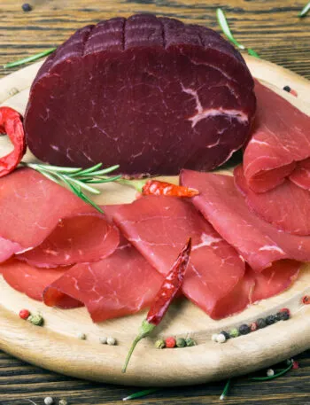 Bresaola affettata e trancio intero su tagliere in legno con rosmarino, pepe e peperoncini, ingrediente leggero e proteico ideale per ricette estive veloci e fit.