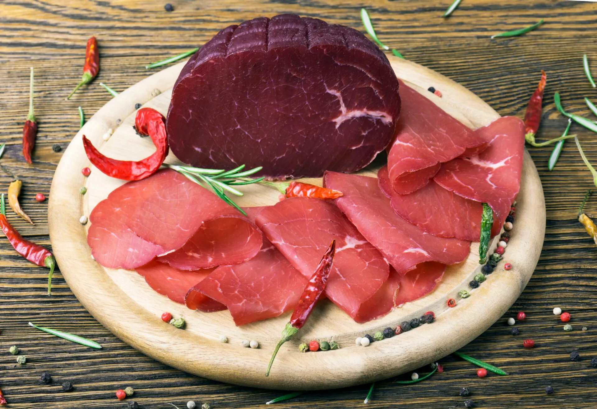 Bresaola affettata e trancio intero su tagliere in legno con rosmarino, pepe e peperoncini, ingrediente leggero e proteico ideale per ricette estive veloci e fit.