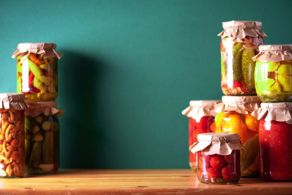 Vasetti di sottaceti e conserve di verdure fatti in casa disposti su un tavolo in legno, con cetrioli, peperoni e pomodori conservati in vetro.