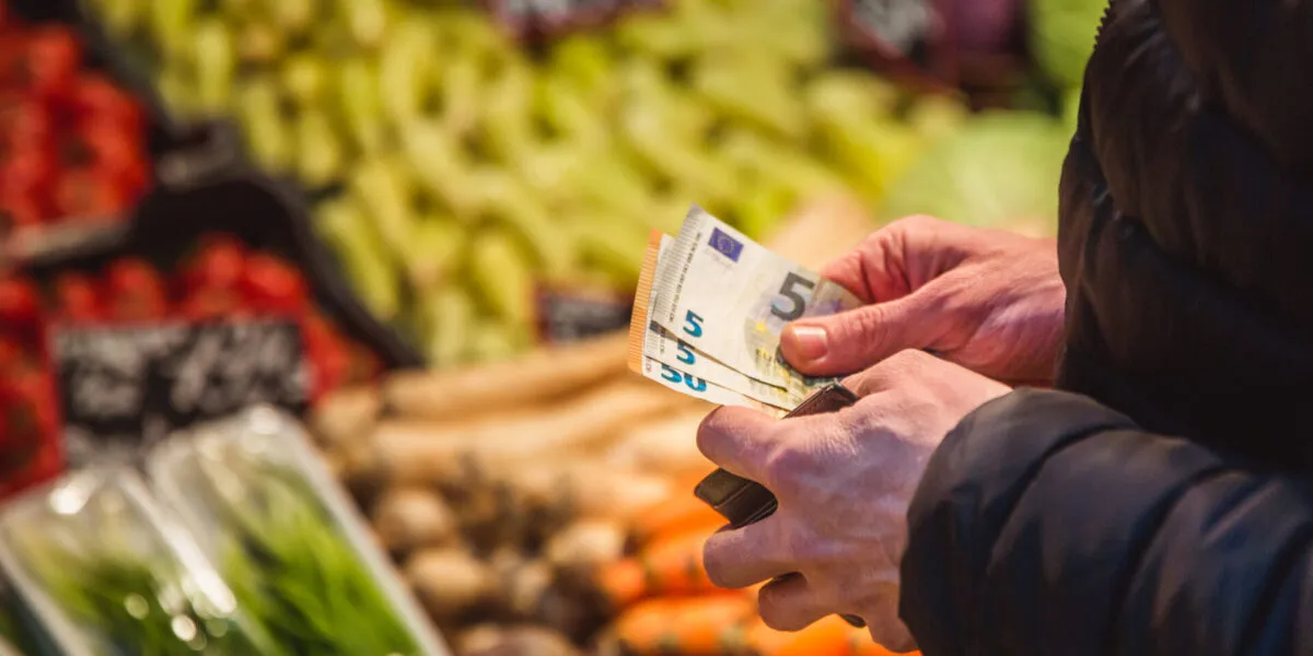 Persona al supermercato mentre conta alcune banconote da 5 euro davanti al reparto ortofrutta, simbolo della gestione del budget e del risparmio sulla spesa settimanale.