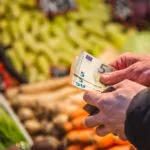 Persona al supermercato mentre conta alcune banconote da 5 euro davanti al reparto ortofrutta, simbolo della gestione del budget e del risparmio sulla spesa settimanale.