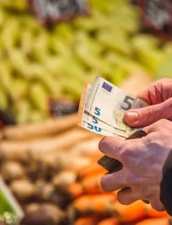 Persona al supermercato mentre conta alcune banconote da 5 euro davanti al reparto ortofrutta, simbolo della gestione del budget e del risparmio sulla spesa settimanale.