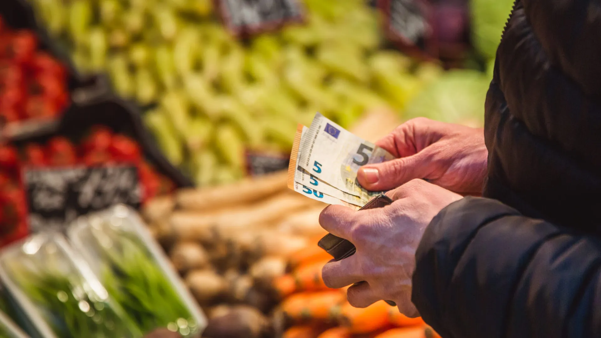 Persona al supermercato mentre conta alcune banconote da 5 euro davanti al reparto ortofrutta, simbolo della gestione del budget e del risparmio sulla spesa settimanale.